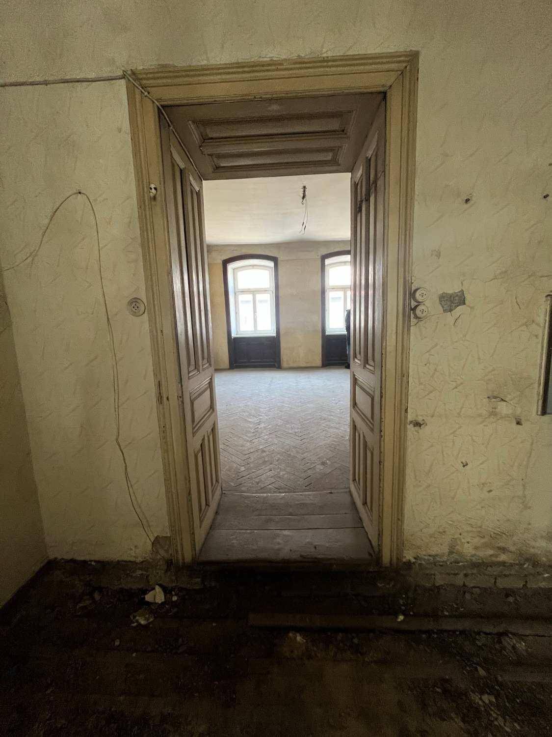 Blick durch eine offene Tür in einen leeren, sanierungsbedürftigen Raum mit zwei Fenstern.