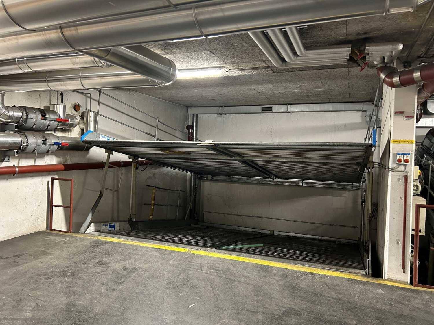 Tiefgarage mit einem mechanischen Parksystem für zwei Fahrzeuge und sichtbaren Versorgungsleitungen an der Decke.