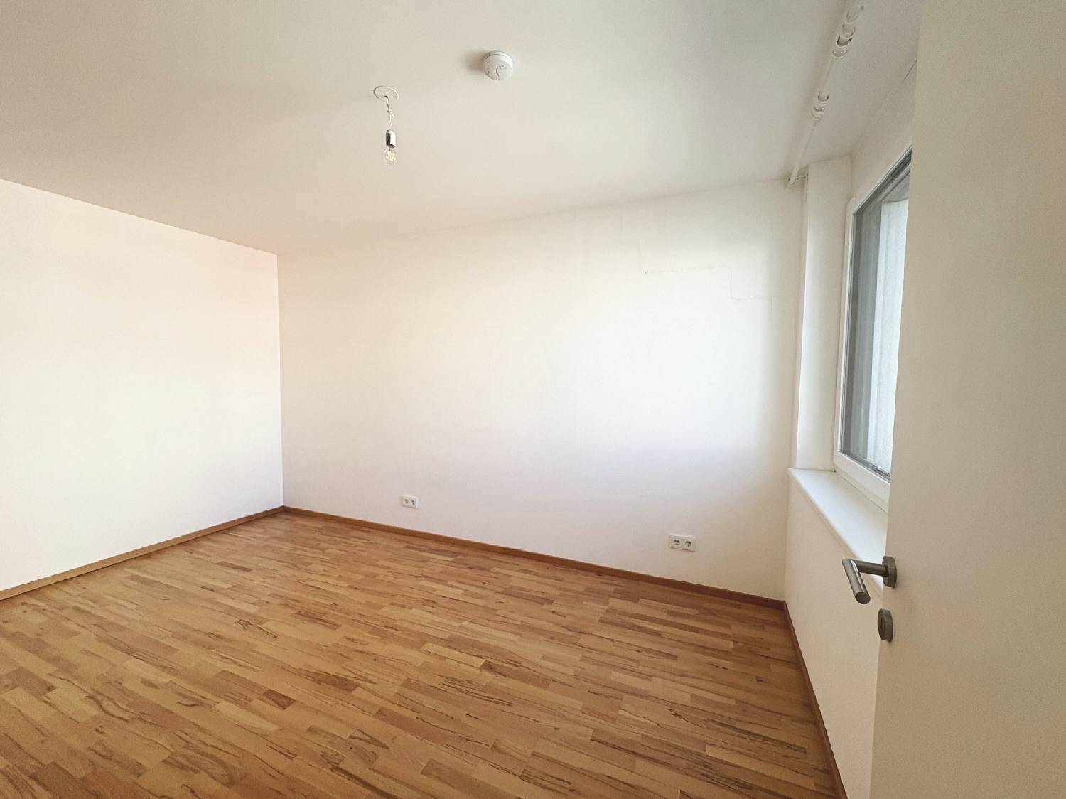 Leeres Zimmer mit Holzboden und einem Fenster, ideal als Schlaf- oder Arbeitszimmer nutzbar.