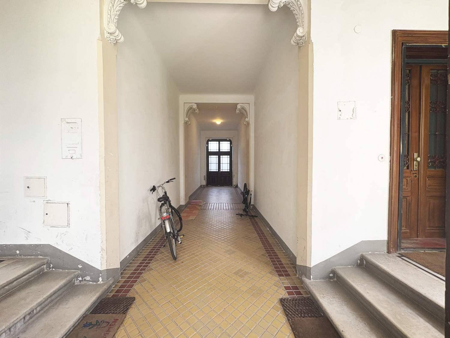 Einfacher Eingangsbereich mit Fliesenboden, weißen Wänden und einer Holztür am Ende des Ganges.