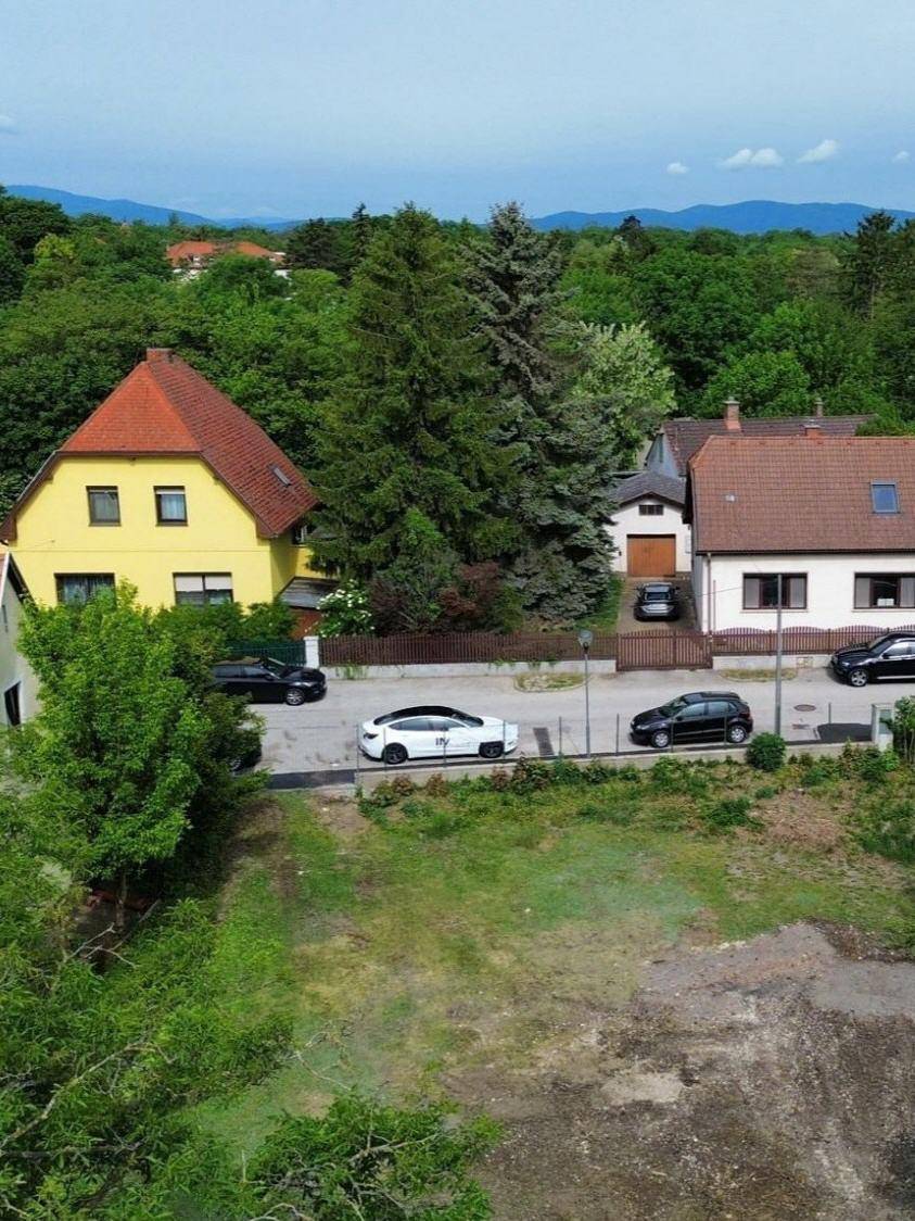 Panoramablick von oben auf das Grundstück, die Straße, Wohnhäuser und eine bewaldete Hügellandschaft im Hintergrund.