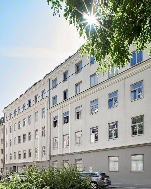 Sonnige Fassade eines modernen Wohngebäudes mit vielen Fenstern und Bäumen im Vordergrund.
