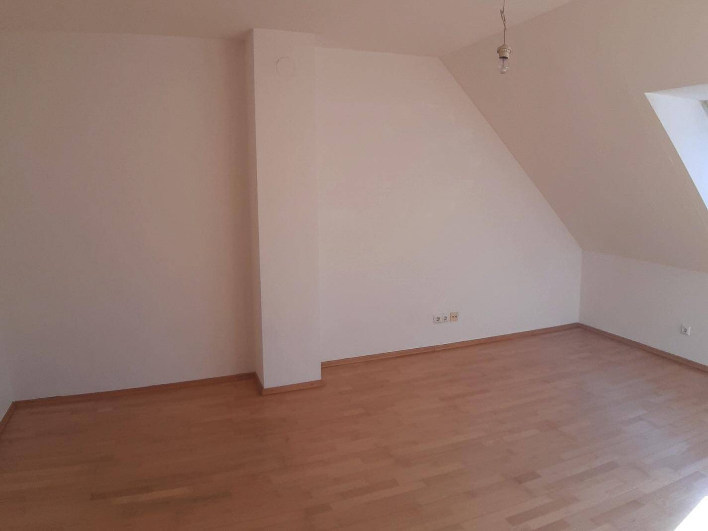 Leerer Raum mit hellem Holzfußboden, weißen Wänden und schräger Dachschräge, durchflutet von Sonnenlicht.
