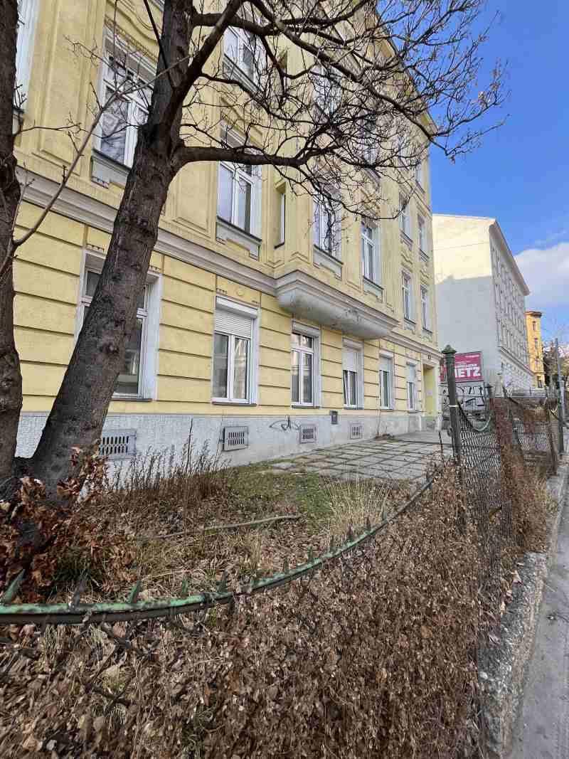 Seitenansicht des Gebäudes von der Straße aus, umgeben von Bäumen und einem Zaun.