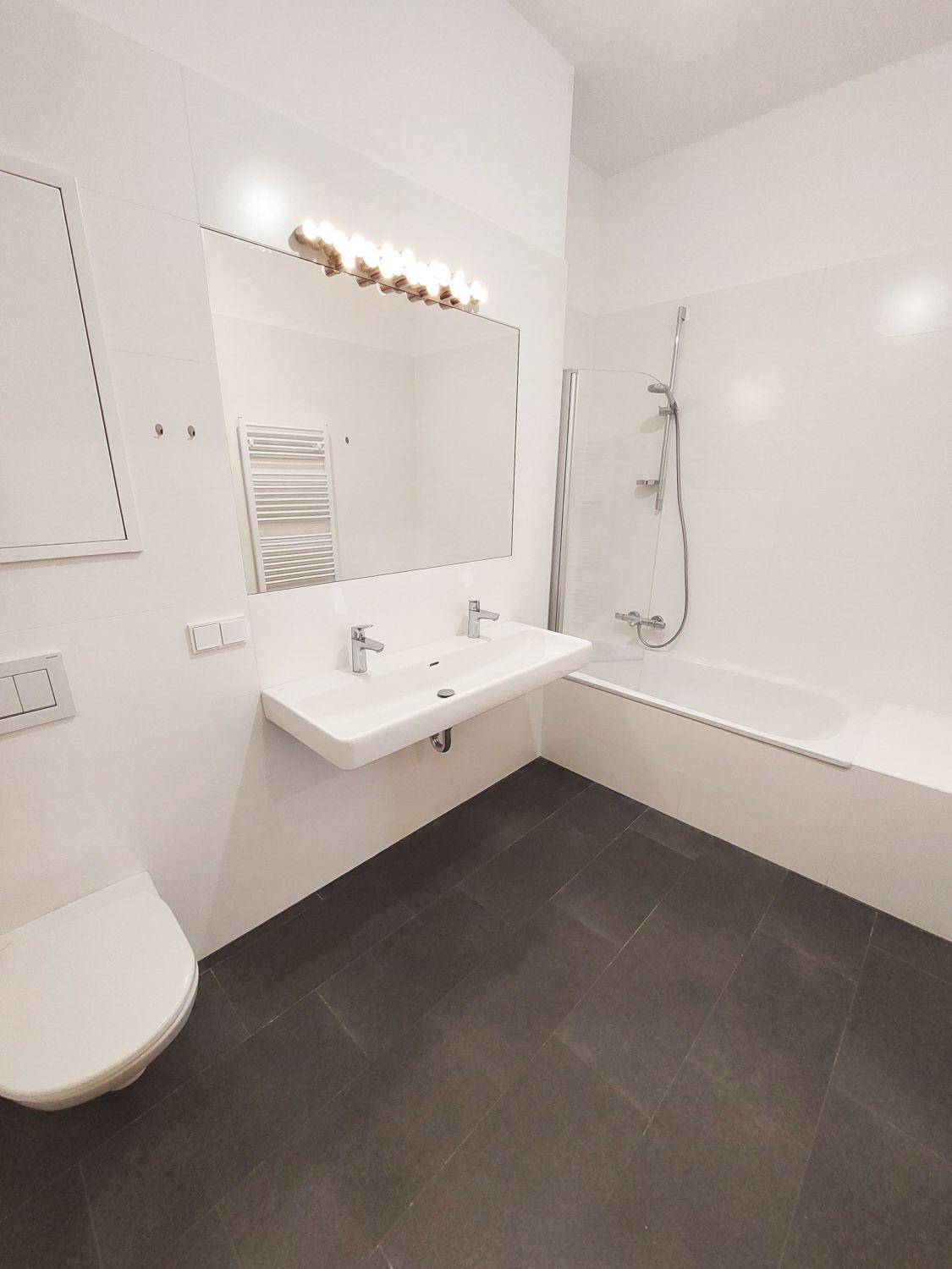 Modernes Badezimmer mit Doppelwaschbecken, Badewanne mit Duschwand, WC und dunklen Fliesen.