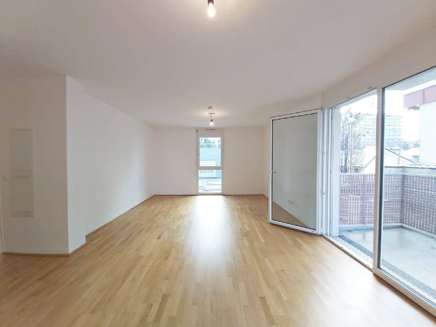 Geräumiges Wohnzimmer mit hellem Parkettboden, großen Fenstern und Zugang zum Außenbereich.