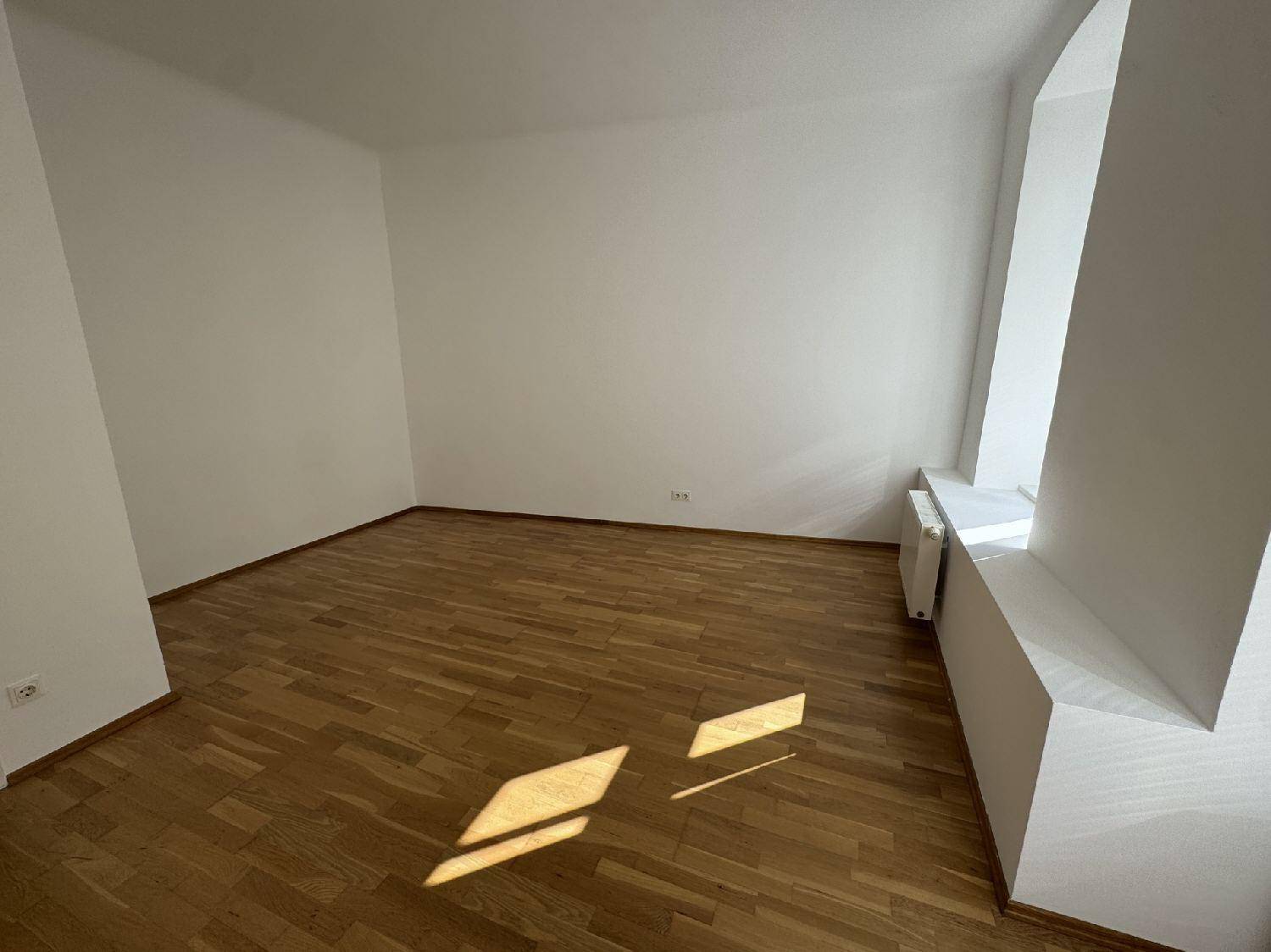 Helles, leeres Zimmer mit Parkettboden und einem Fenster, das viel Sonnenlicht hereinlässt.