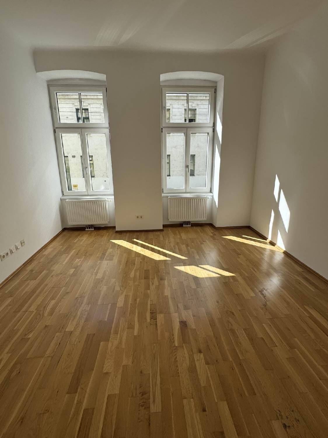 Lichtdurchflutetes Zimmer mit Holzboden und zwei hohen Fenstern, die den Raum erhellen.