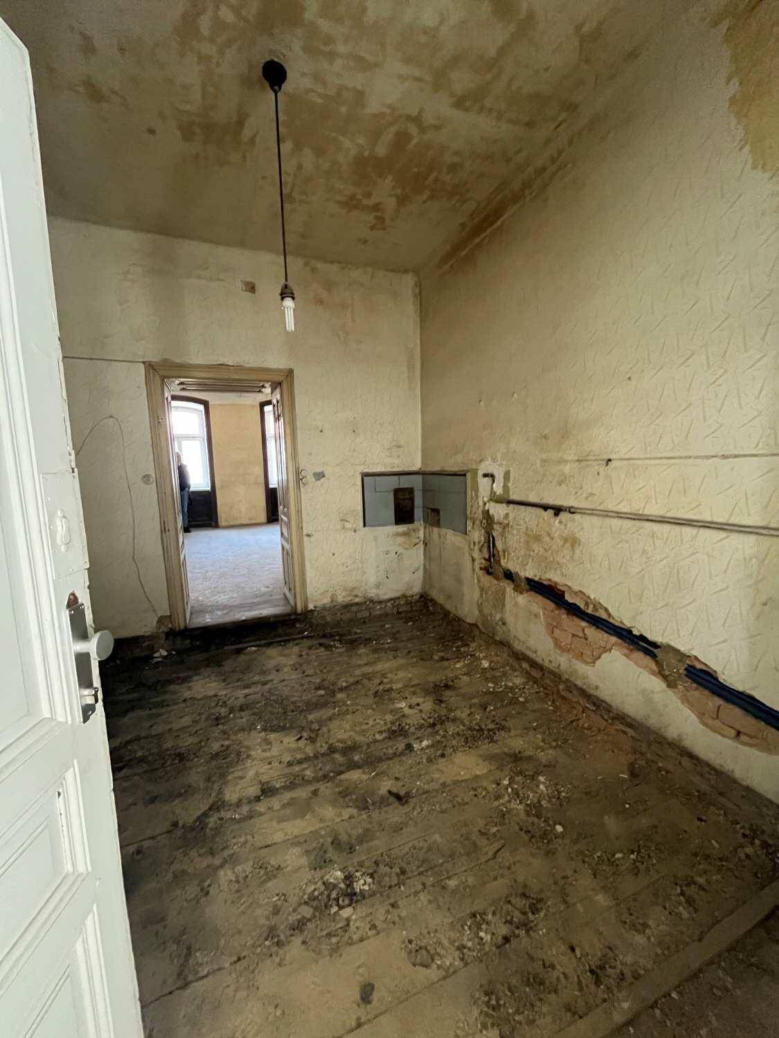 Sanierungsbedürftige Küche mit freiliegenden Rohren, abgenutztem Holzboden und beschädigten Wänden.