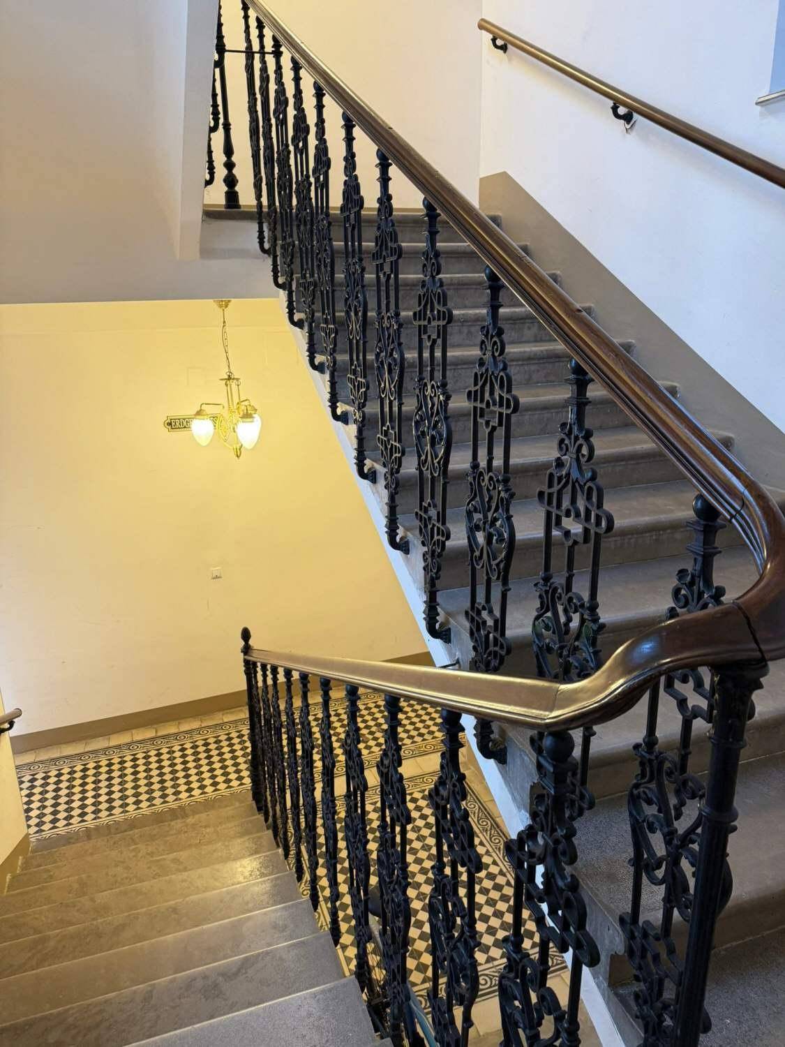Großzügiges Treppenhaus mit kunstvollem schmiedeeisernem Geländer und Schachbrettmuster-Fliesenboden.