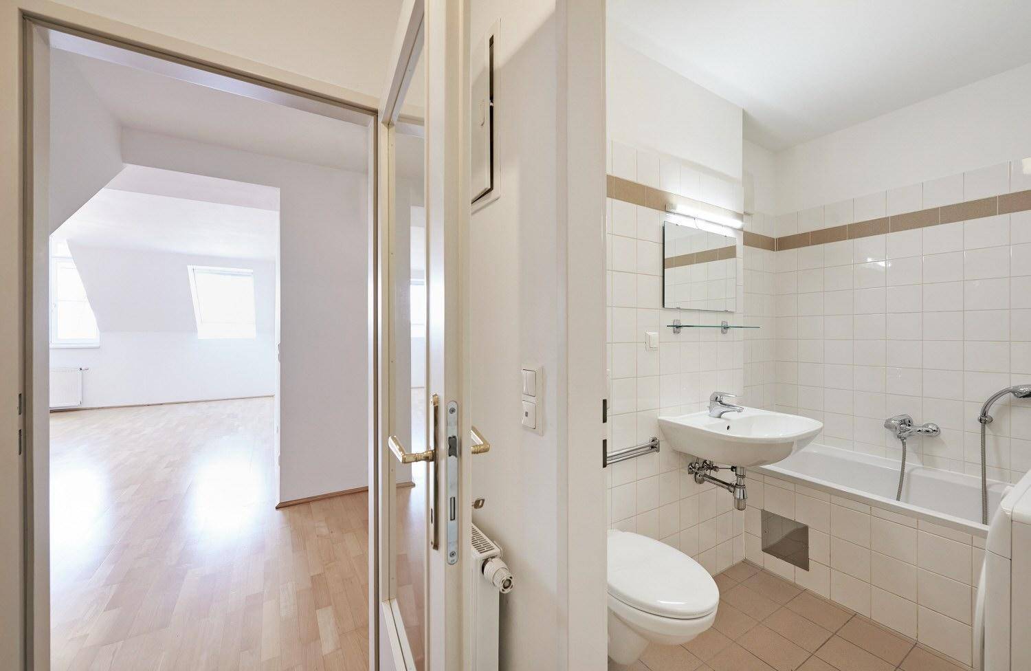 Badezimmer mit Badewanne, WC und Waschbecken, gefliest in Weiß mit braunem Akzentstreifen.