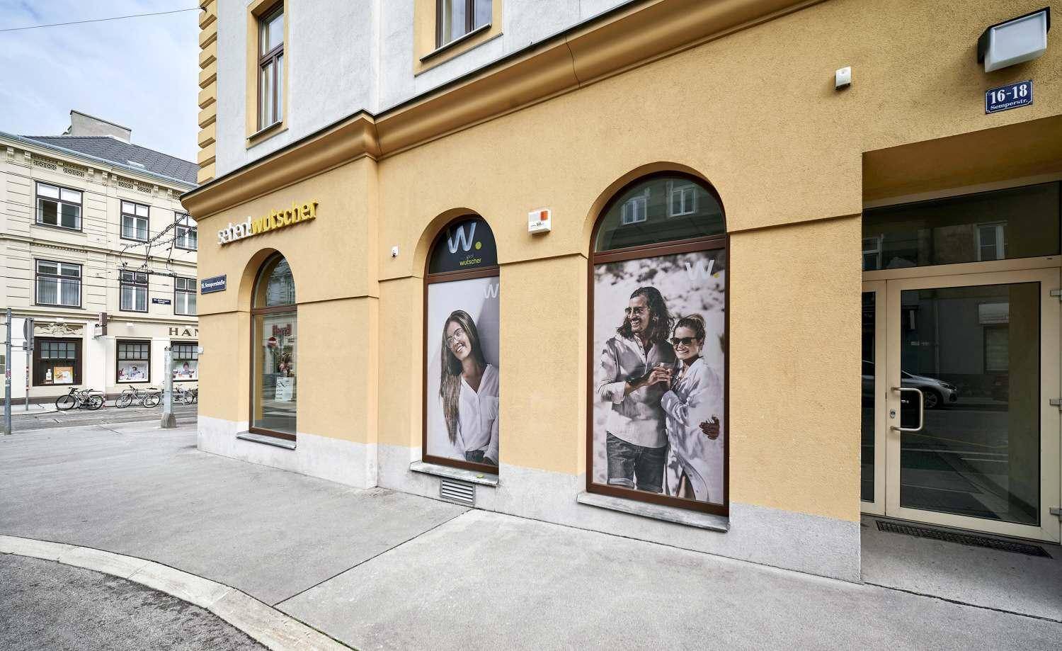 Eckansicht der Gebäudefassade mit Schaufenstern und Passanten auf dem Bürgersteig in einer städtischen Umgebung.