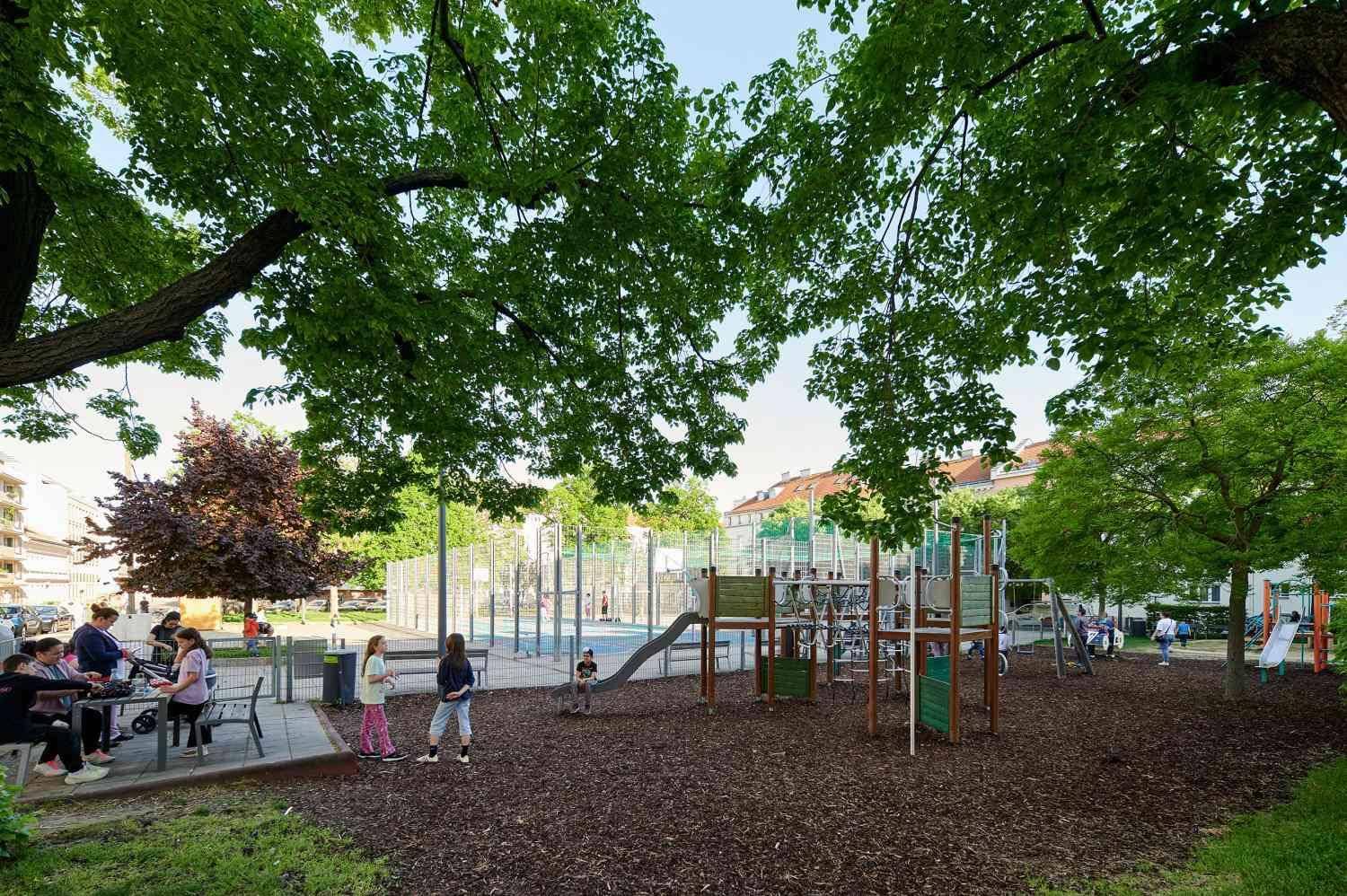 Belebtes öffentliches Spielplatzgelände mit Kindern, Bäumen und Spielgeräten in einer Grünanlage.