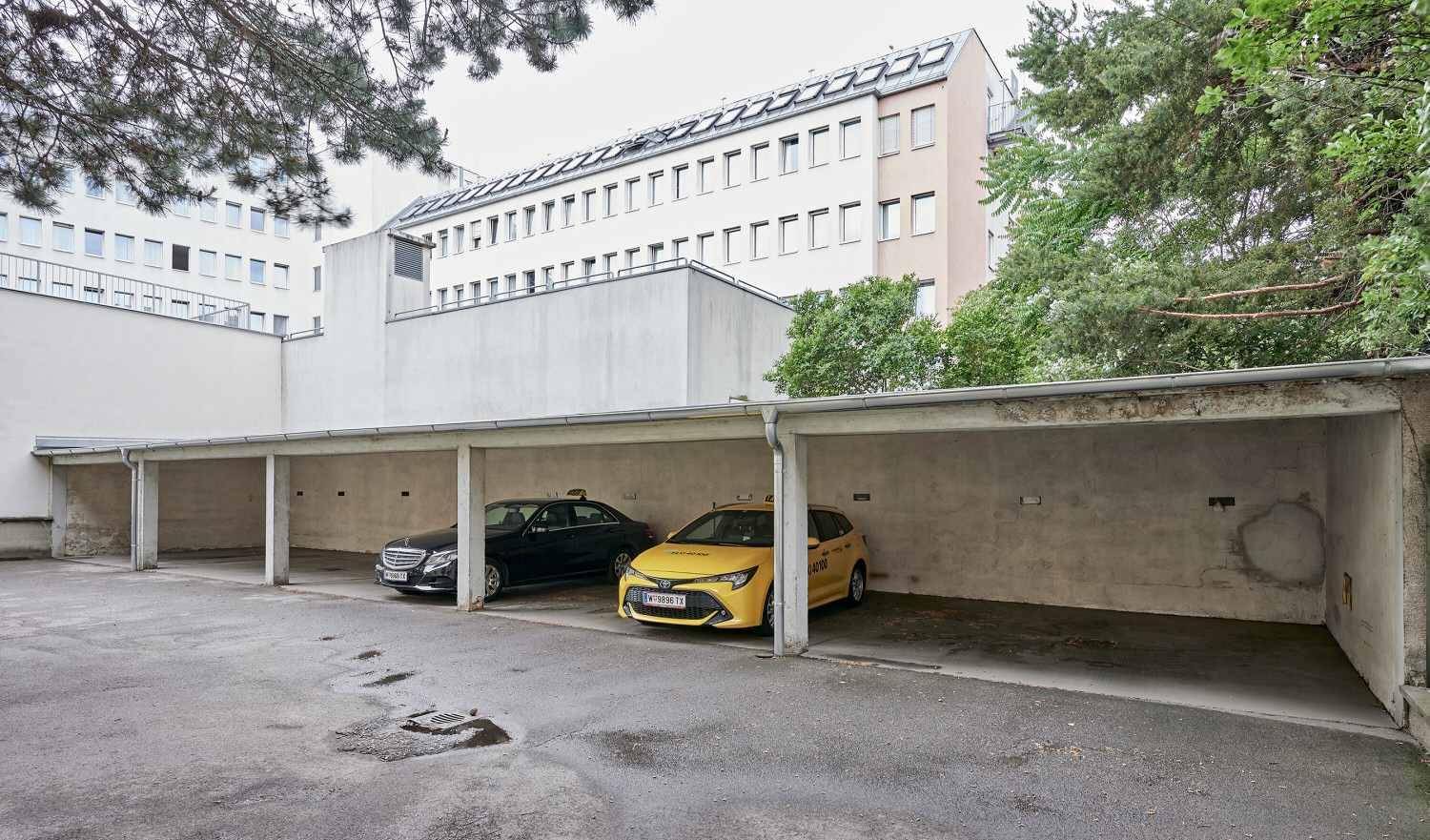 Überdachte Parkplätze im Freien mit zwei geparkten Autos und Bäumen im Hintergrund.