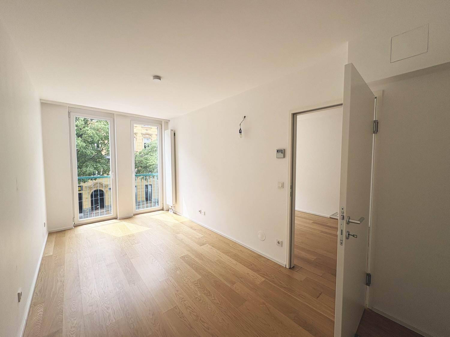 Lichtdurchfluteter Wohnbereich mit Holzboden, französischen Balkonen und offener Tür, die viel Helligkeit hereinlässt.