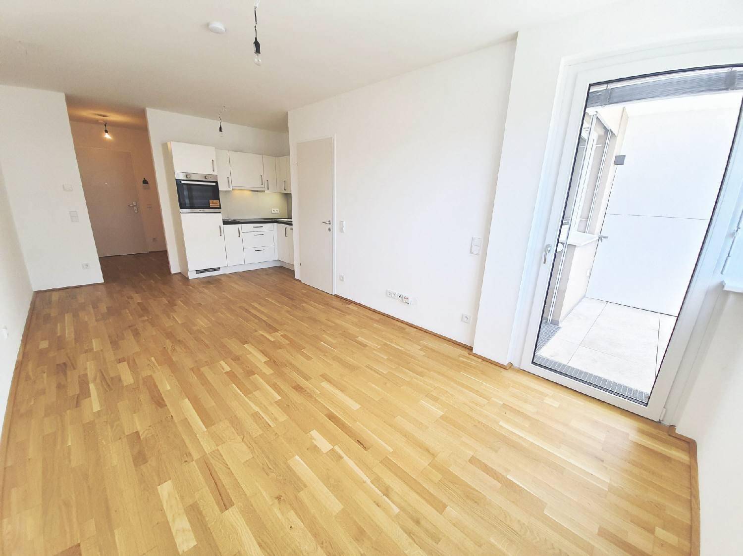 Geräumiges Wohnzimmer mit offener Küchenzeile, hellem Parkettboden und direktem Balkonzugang.