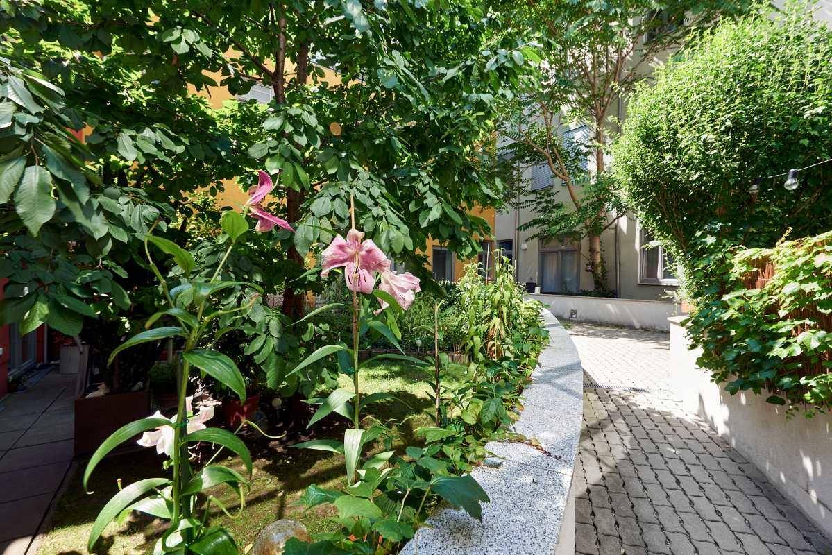 Grüner Innenhof mit blühenden Pflanzen und einem gepflasterten Weg, umgeben von Gebäuden.