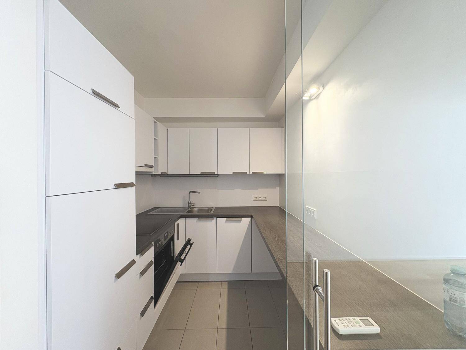 Moderne Einbauküche mit weißen Schränken, dunkler Arbeitsplatte und integriertem Backofen, durch Glasschiebetür abgetrennt.