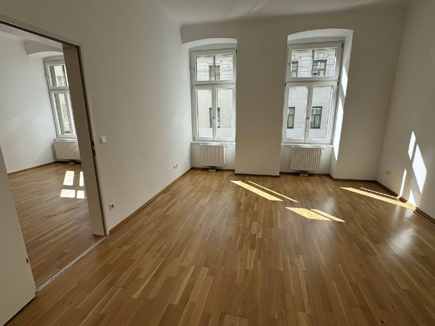 Geräumiges, helles Zimmer mit Parkettboden und zwei Fenstern, das in einen weiteren Raum führt.