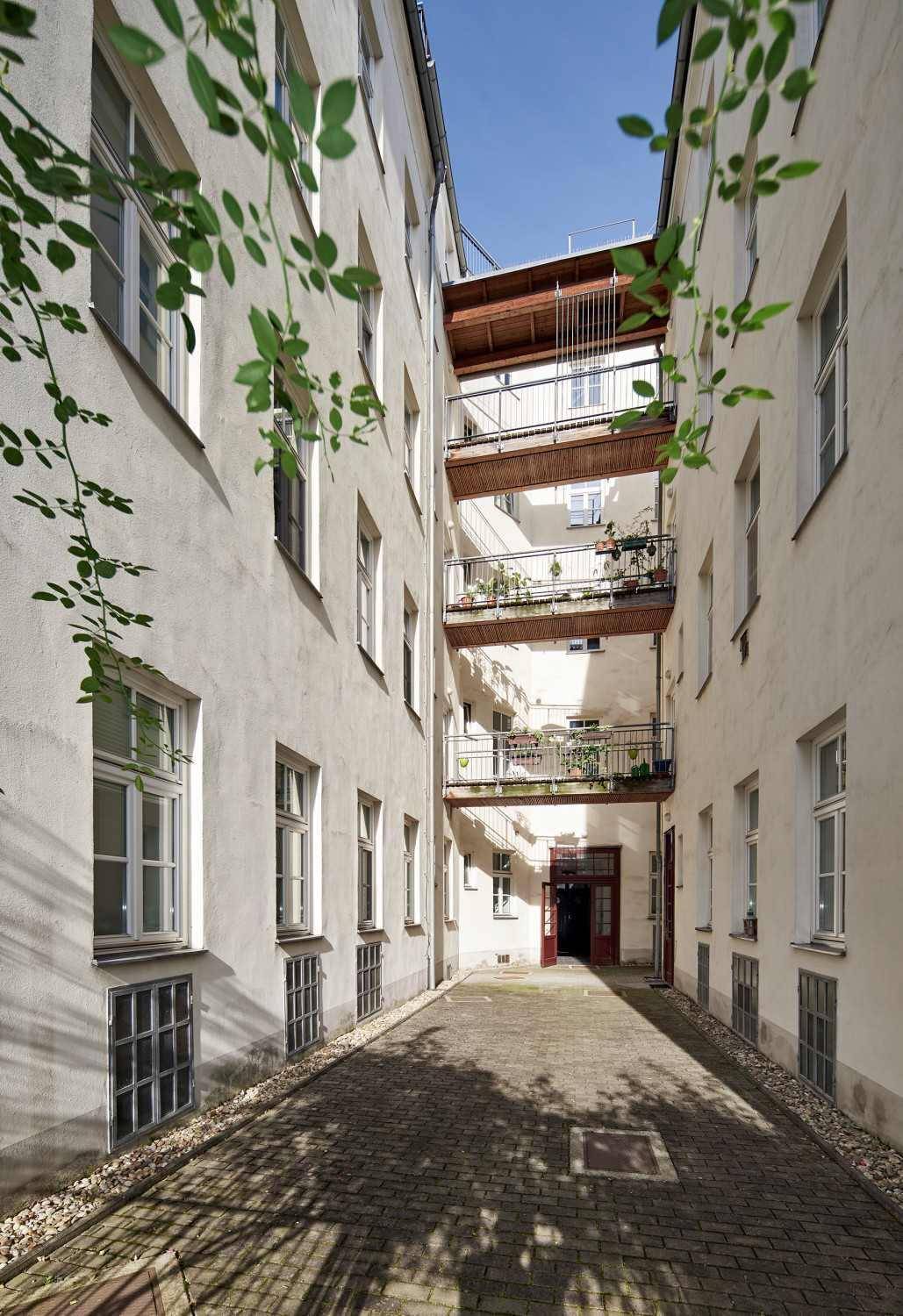 Heller Innenhof mit mehreren Balkonen und einer Holzkonstruktion, umgeben von Gebäuden und etwas Grün.