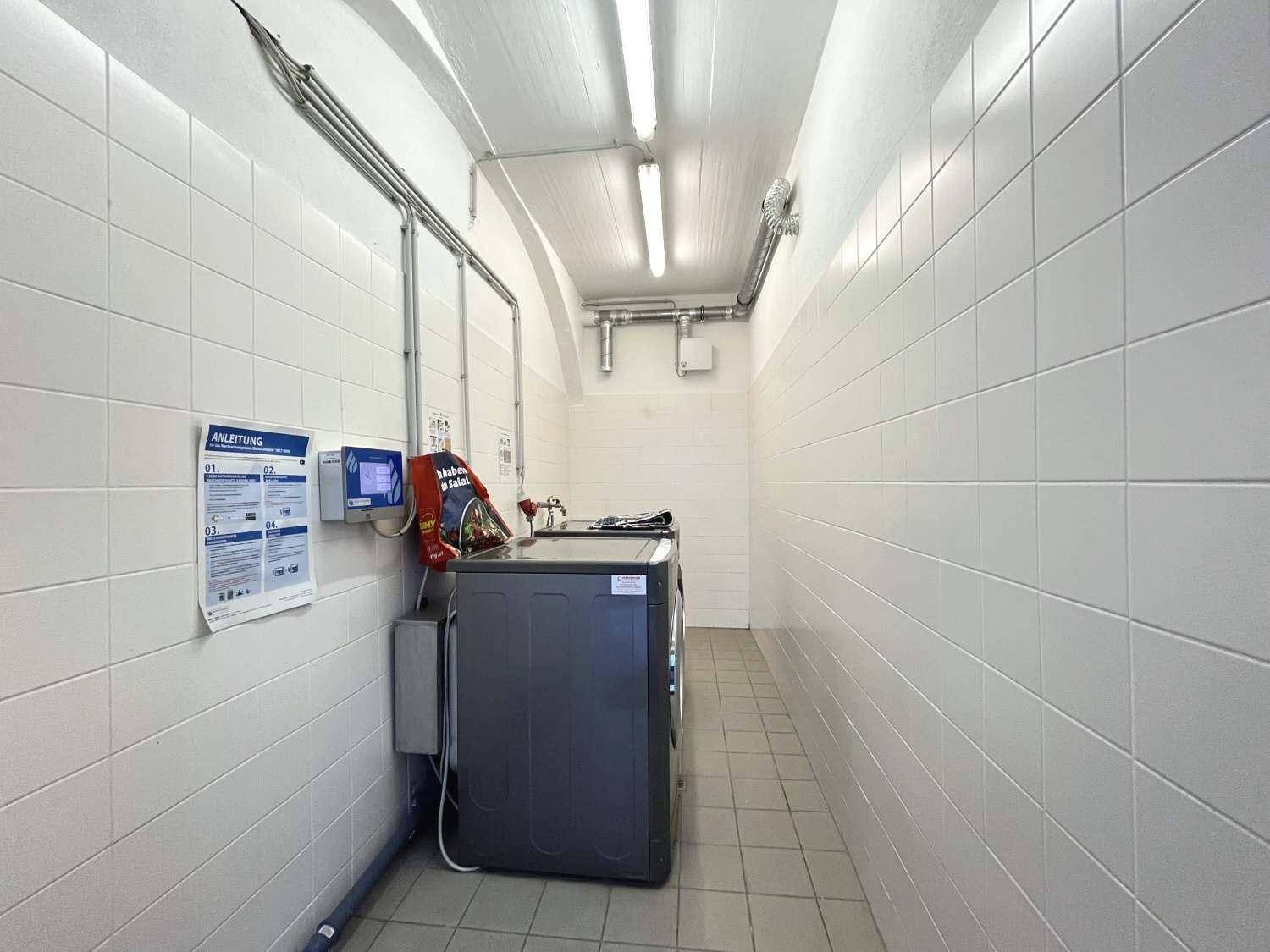 Gemeinschaftlicher Waschraum im Keller mit einer Waschmaschine und weiß gefliesten Wänden.