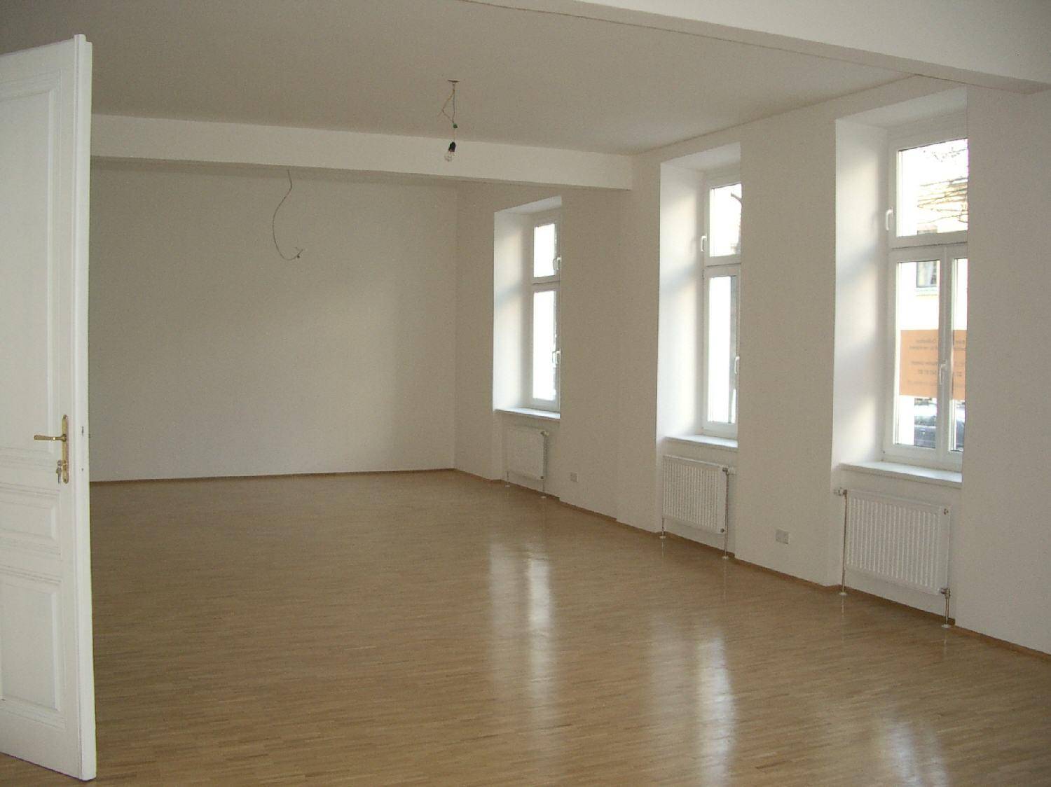 Geräumiger, heller Wohnraum mit Holzboden und großen Fenstern, die viel Tageslicht hereinlassen.