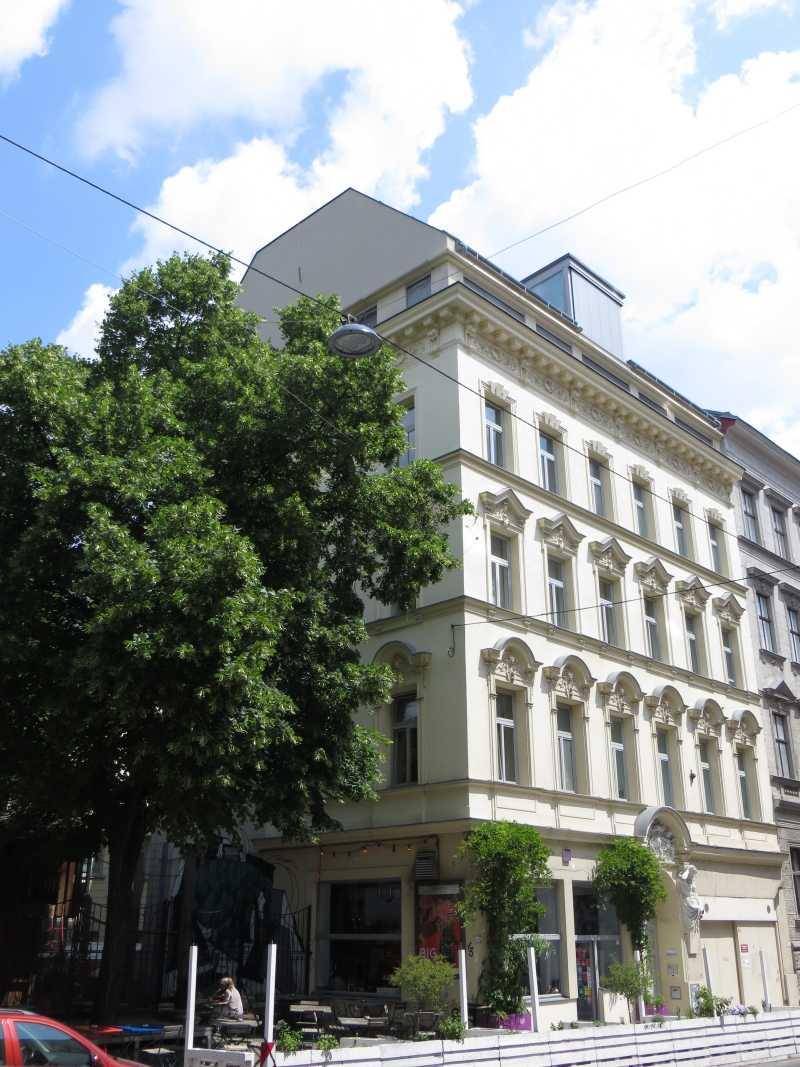 Historisches Gebäude mit reich verzierter Fassade und großen Bäumen davor an einem sonnigen Tag.
