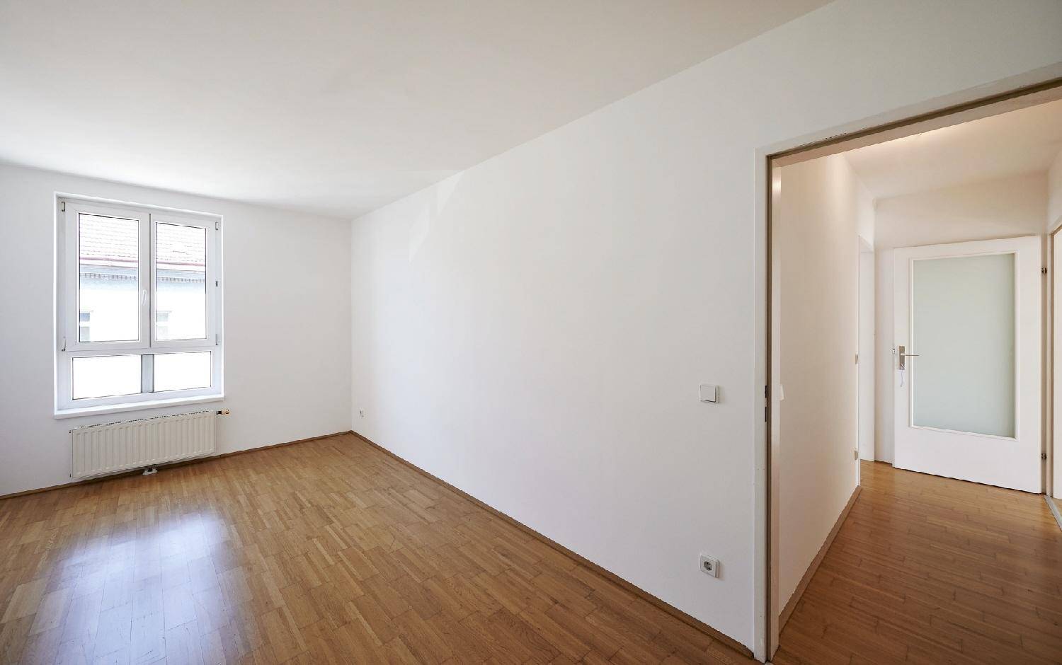 Geräumiges Zimmer mit Holzboden, weißen Wänden und einem Fenster mit Blick nach draußen.