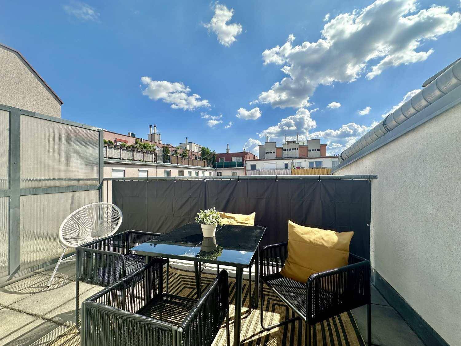Sonniger Balkon mit modernen Outdoor-Möbeln und Blick auf die umliegenden Gebäude und den Himmel.