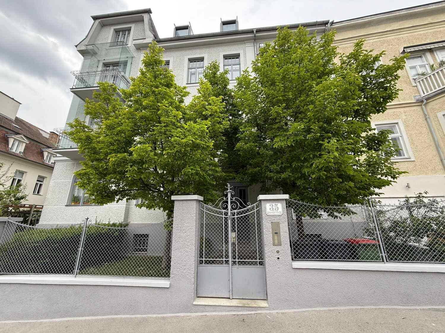 Elegante Fassade des Gebäudes mit grünen Bäumen und einem schmiedeeisernen Eingangstor.