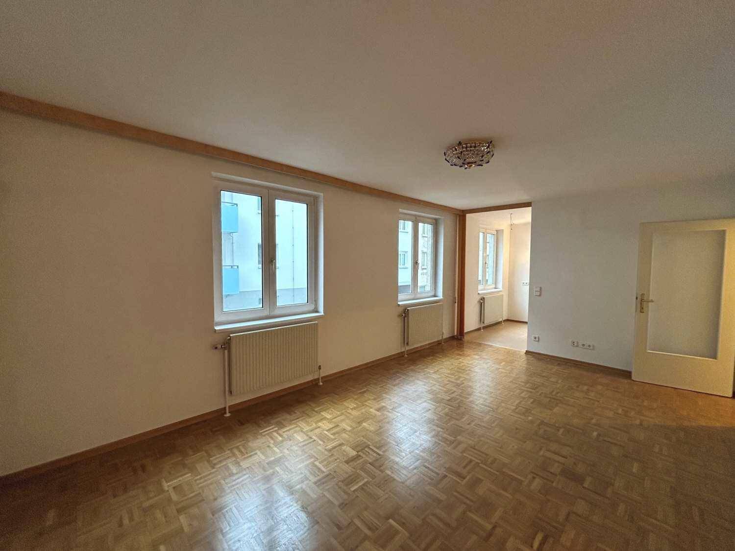 Helles, leeres Wohnzimmer mit Parkettboden und zwei großen Fenstern, die viel Tageslicht hereinlassen.