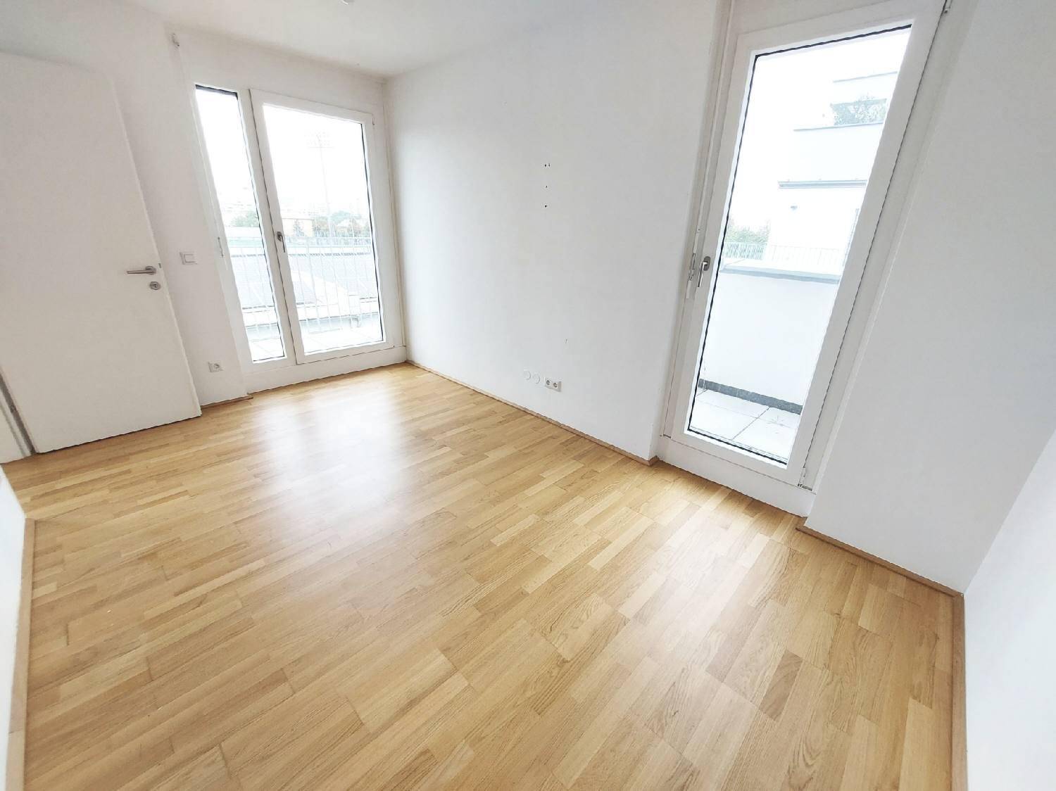 Helles Zimmer mit Parkettboden und Zugang zu einem Außenbereich durch große Türen.