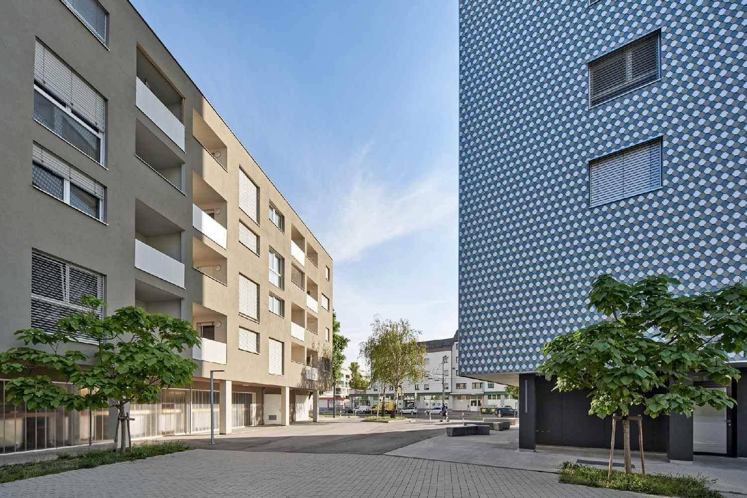 Zwei moderne Wohngebäude mit unterschiedlichen Fassaden und gepflastertem Vorplatz.