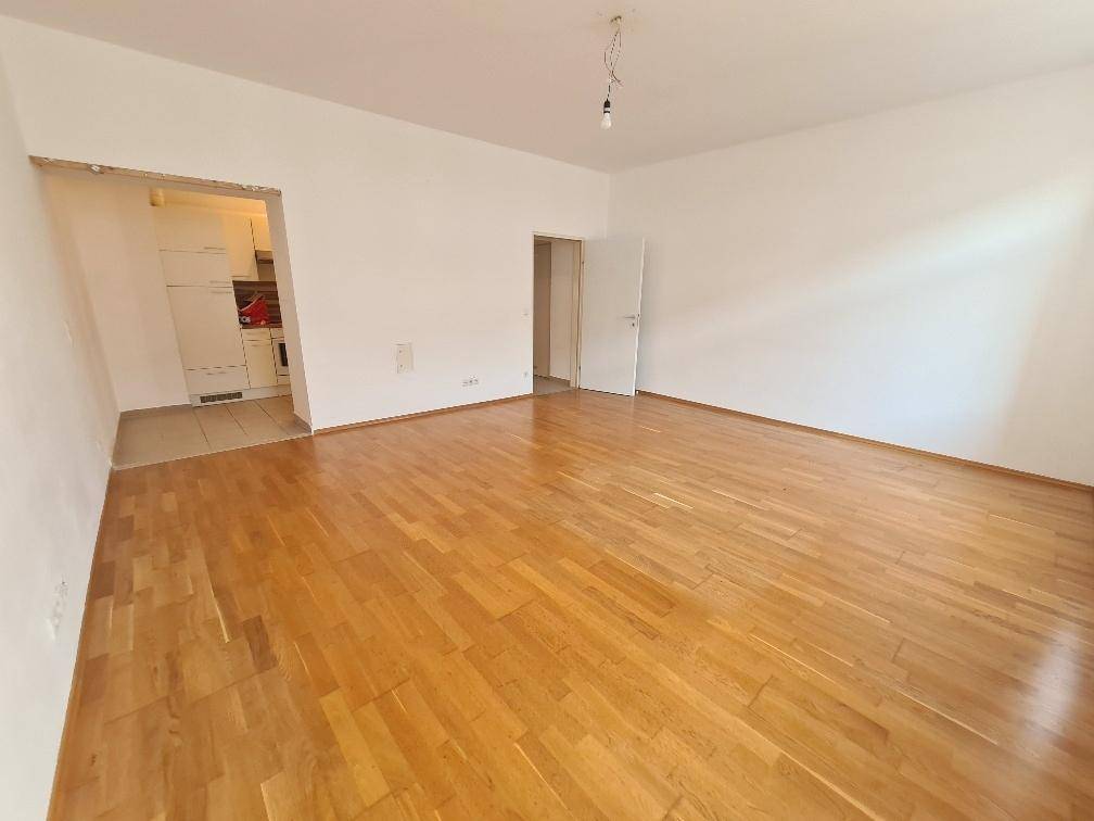 Geräumiges, leeres Zimmer mit Parkettboden und Zugang zu einem Kochbereich.