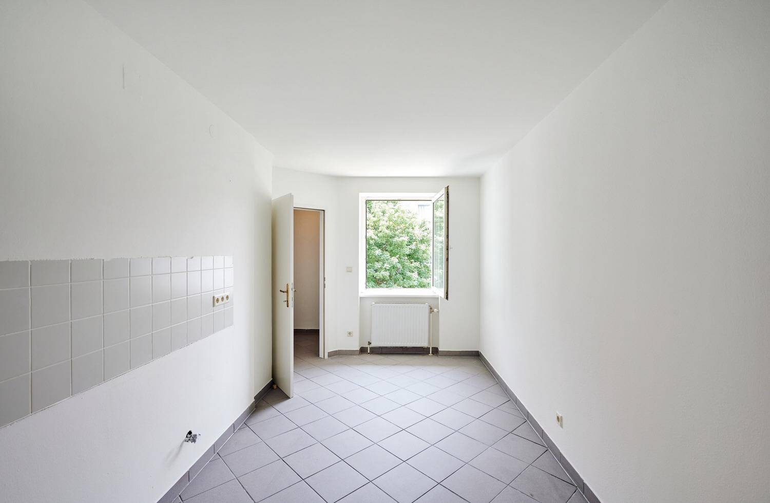 Leere Küche mit grauen Fliesen, weiß gestrichenen Wänden und einem Fenster mit Blick ins Grüne.