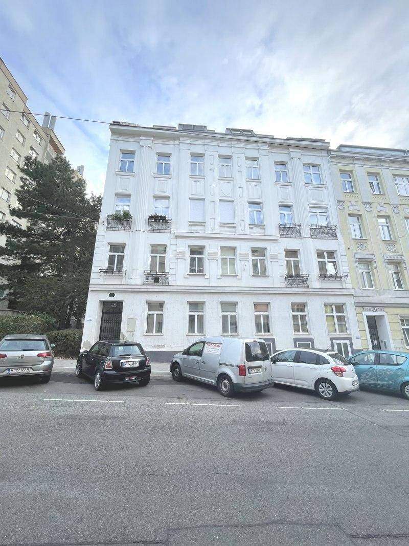 Weißes Mehrfamilienhaus mit parkenden Autos auf der Straße unter bewölktem Himmel.