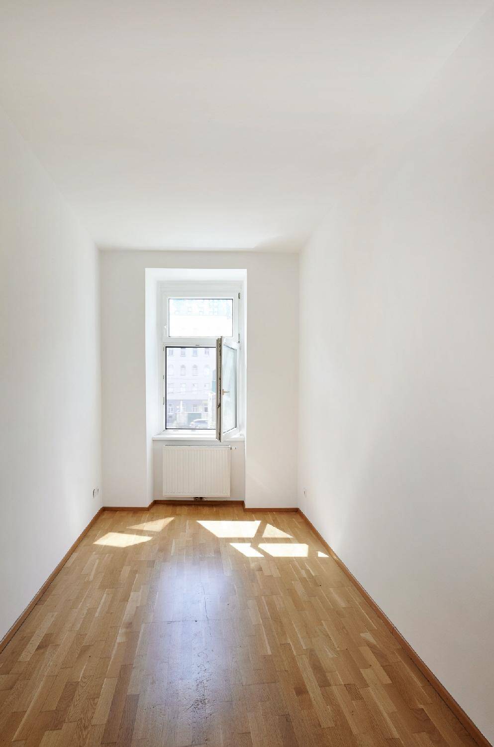 Kleiner, heller Raum mit Parkettboden und einem Fenster, das viel Sonnenlicht hereinlässt.