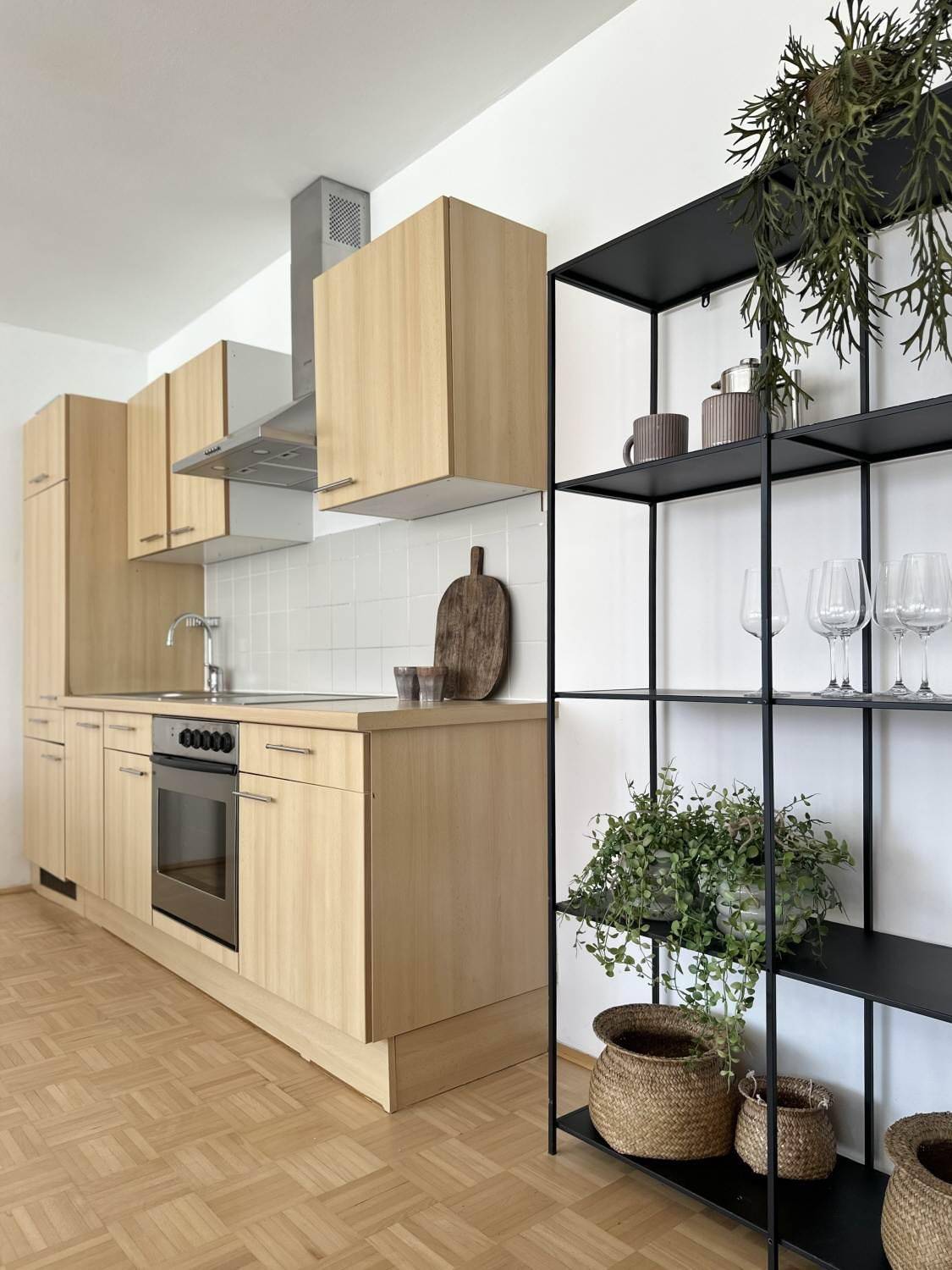 Moderne Einbauküche mit hellen Holzfronten, integrierten Geräten und offenem Regal mit Pflanzen.