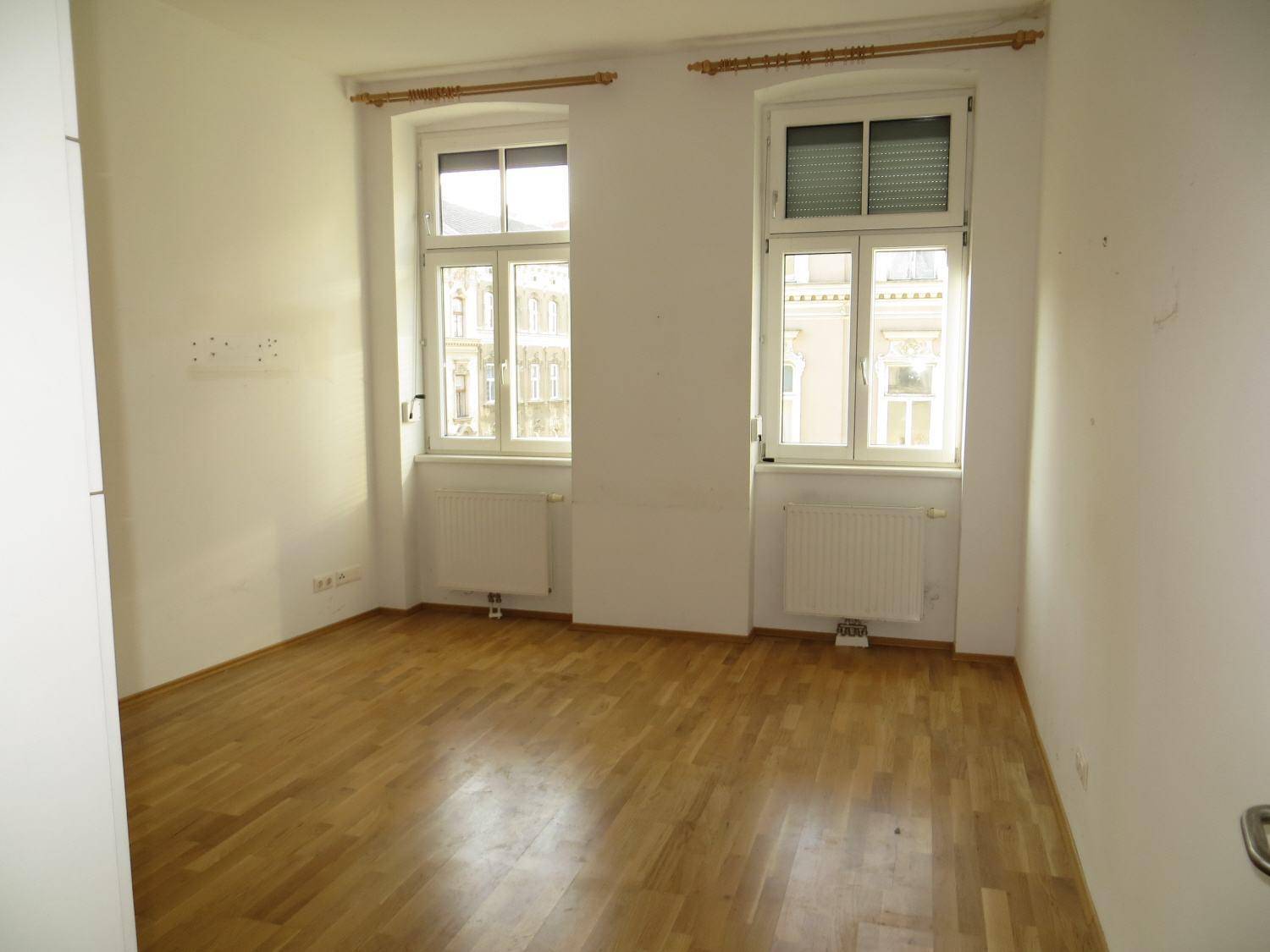 Leeres Zimmer mit zwei großen Fenstern, Heizkörpern und hellem Parkettboden.