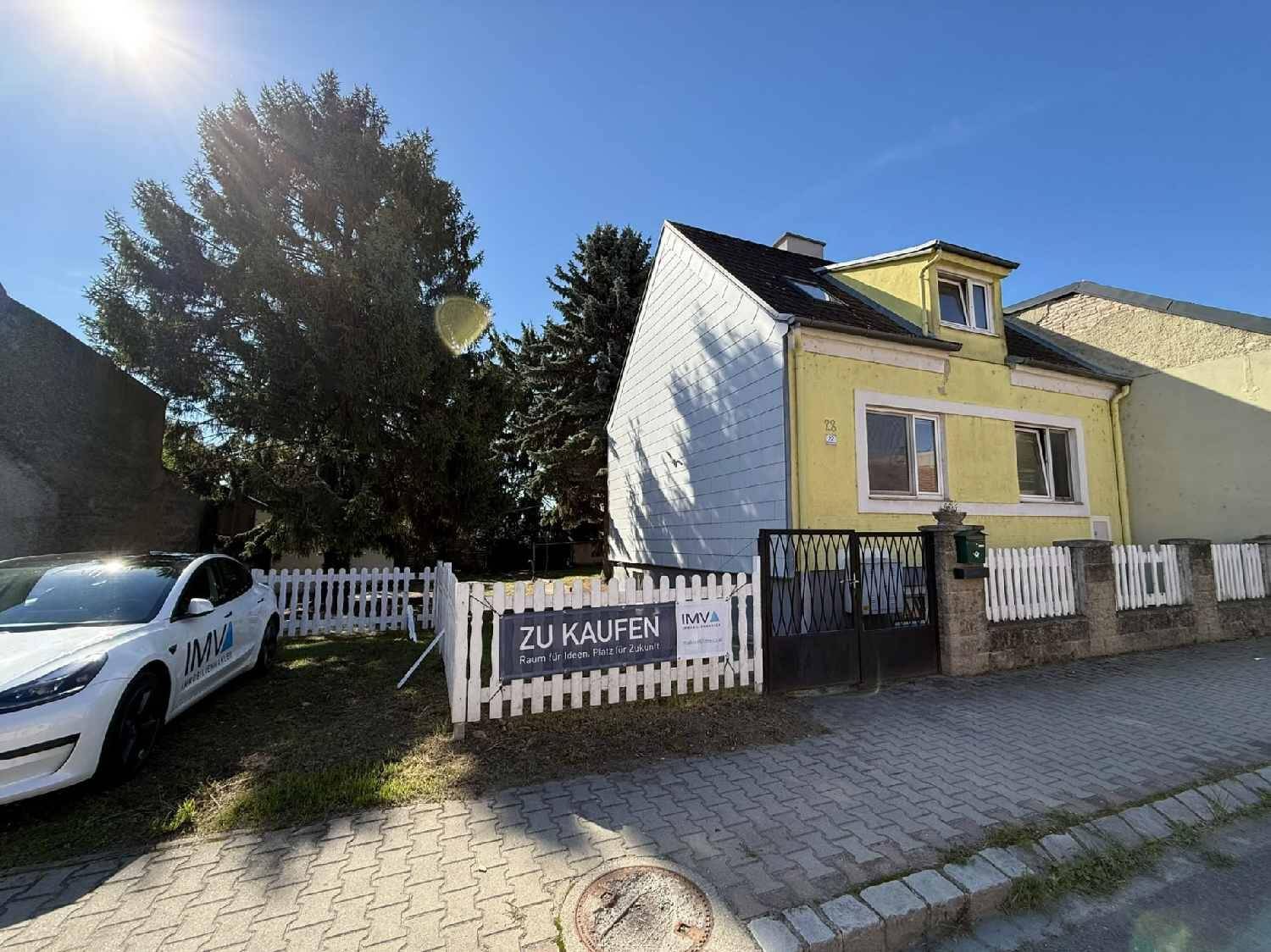 Gelbes Einfamilienhaus mit weißem Zaun und 'ZU KAUFEN' Schild an einem sonnigen Tag.