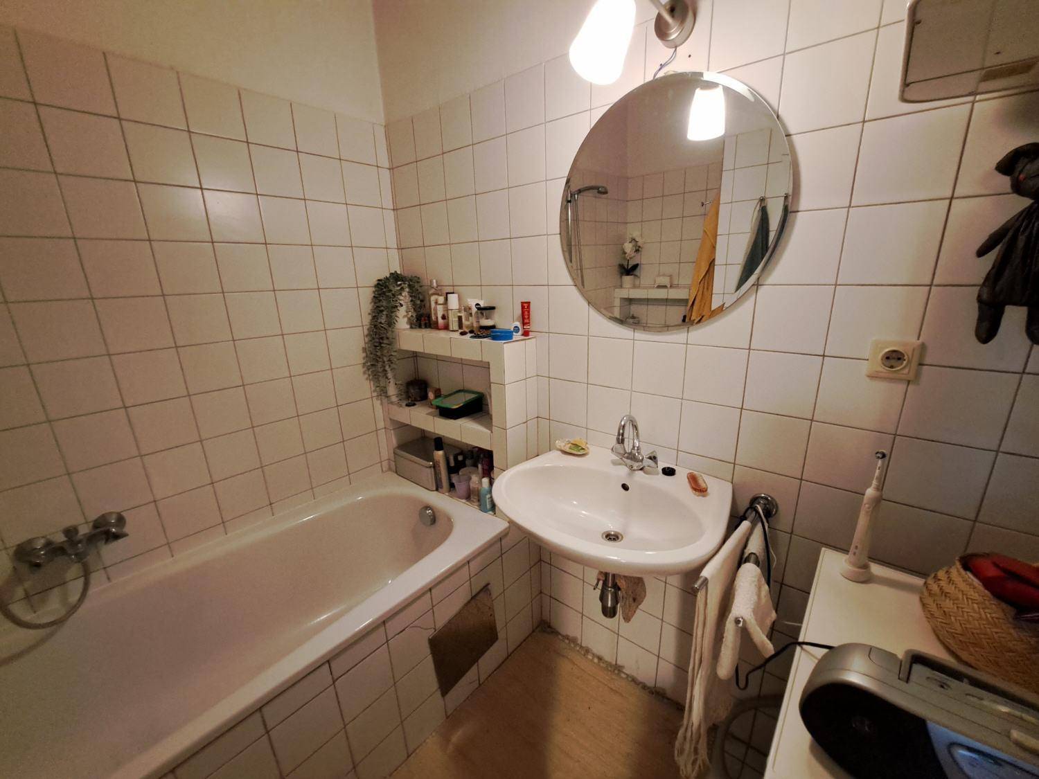 Badezimmer mit weißen Fliesen, Badewanne, Waschbecken und einem runden Spiegel, funktional und sauber.
