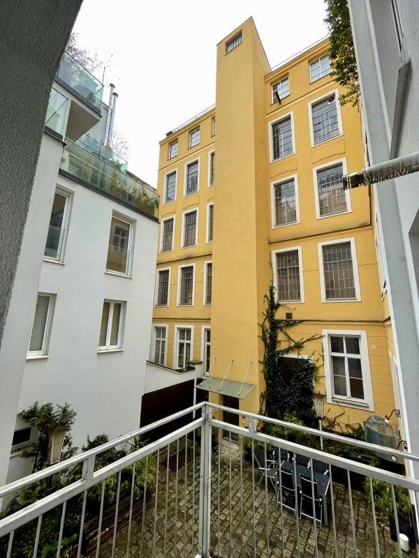 Blick von einem Balkon auf einen begrünten Innenhof mit gelber Gebäudefassade und Sitzgelegenheiten.