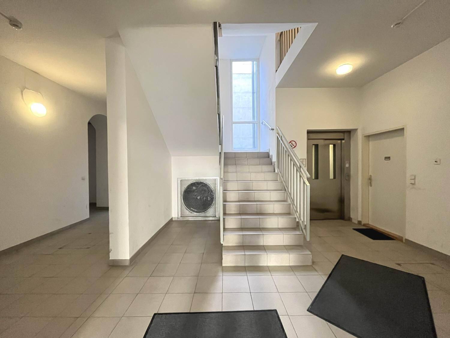 Heller Eingangsbereich mit Treppenhaus, Fliesenboden und einem Aufzug im Hintergrund des Gebäudes.