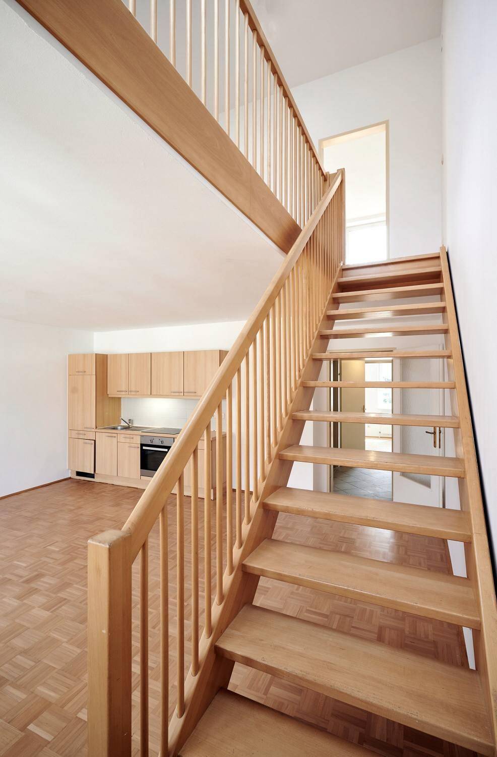 Blick von der Holztreppe in den offenen Wohnbereich mit integrierter Küchenzeile und Parkettboden.