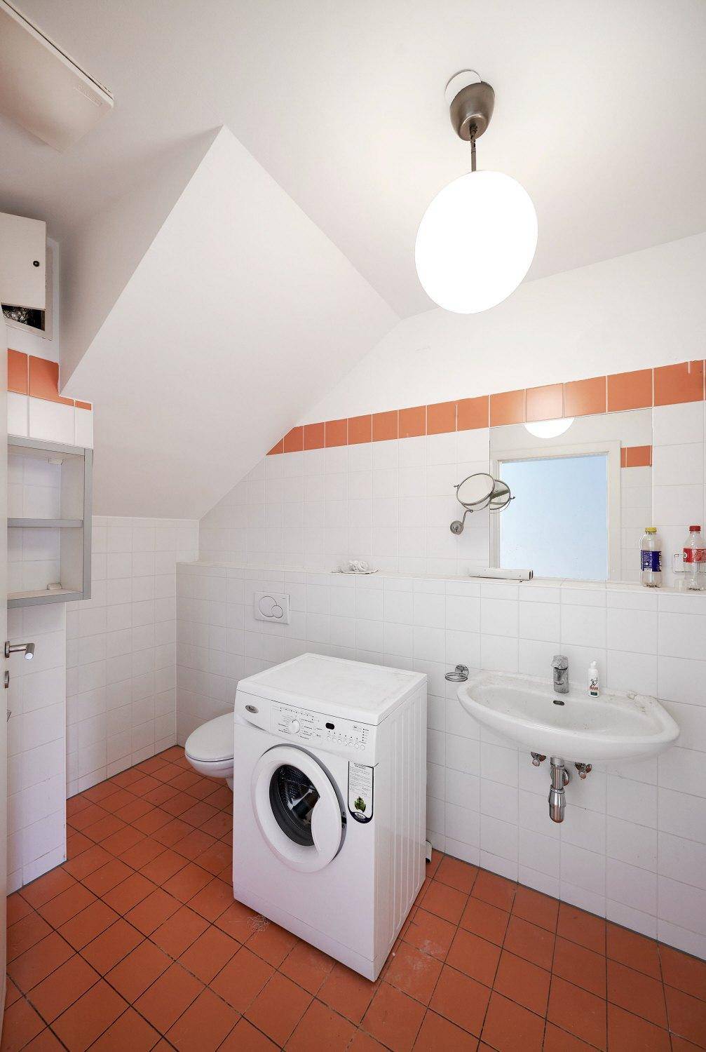 Badezimmer mit Waschmaschine, Waschbecken, WC und orangefarbenen Akzentfliesen.