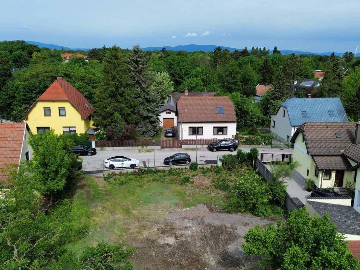 Panoramablick von oben auf das Grundstück, die Straße, Wohnhäuser und eine bewaldete Hügellandschaft im Hintergrund.