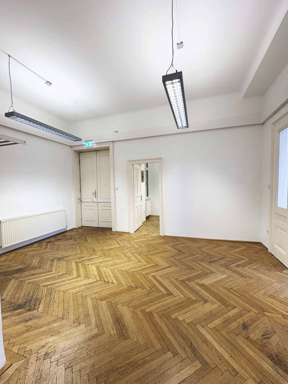 Geräumiges Zimmer mit Fischgrätparkett, Stuckdecke und hohen Fenstern, bietet viel Tageslicht.