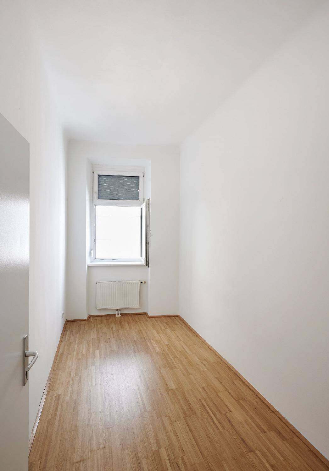 Schmaler, heller Raum mit Parkettboden und einem geöffneten Fenster, ideal als Schlafzimmer.