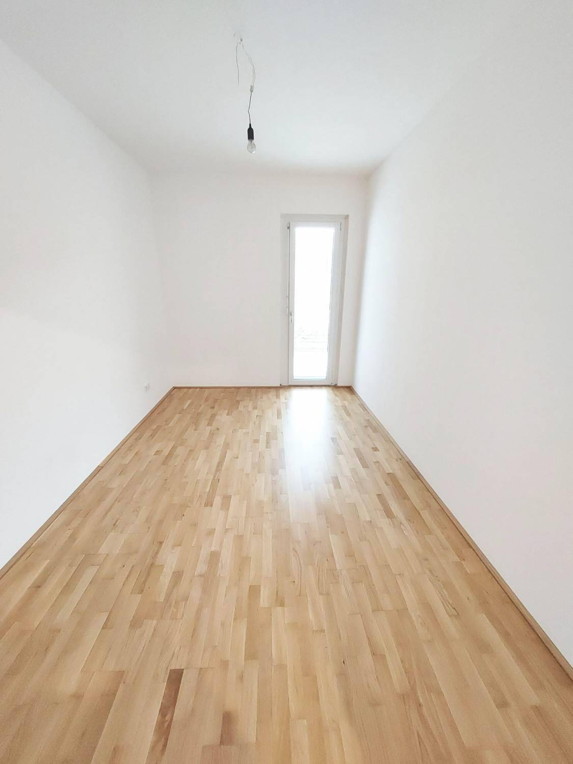 Langes, helles Zimmer mit Parkettboden, weißen Wänden und einer Tür, die zu einem Außenbereich führt.