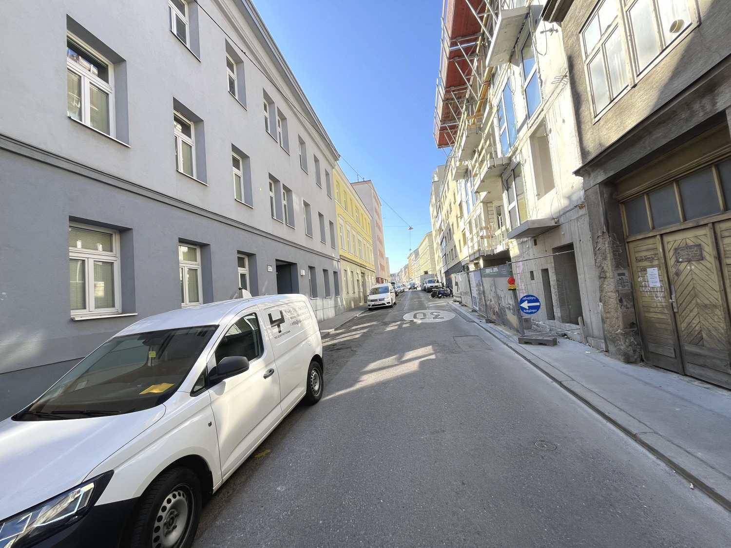 Blick auf eine städtische Straße mit parkenden Fahrzeugen und Gebäuden auf beiden Seiten unter blauem Himmel.